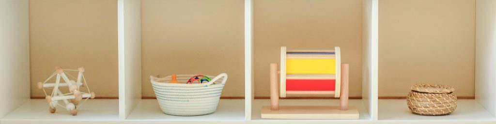Montessori shelf with four sections. Toys left to right: skwish rattle, basket of balls, spinning drum, small wicker basket with lid