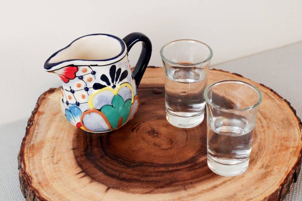 Wooden trivet with a colorful small pitcher of water and 2 small shot glasses with water