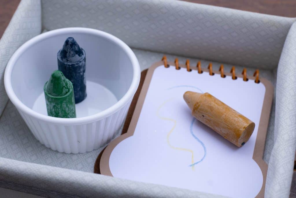 Tray with a dish of 2 beeswax crayons and a small notepad with scribbles on it and a crayon sitting on top of it. 