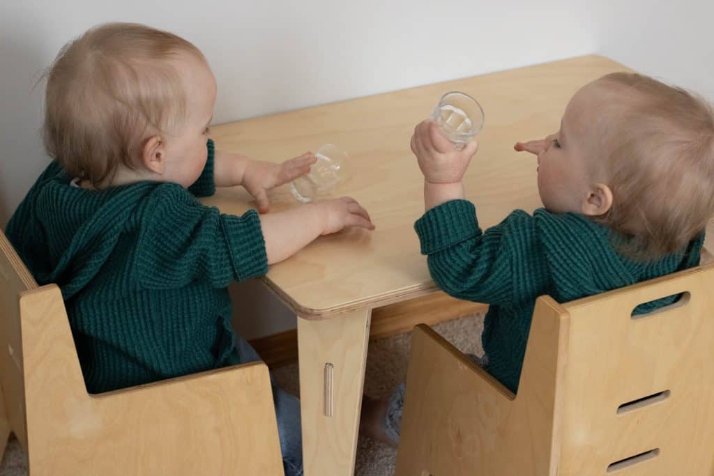Twins sitting at a small Montessori weaning table in chairs holding cups