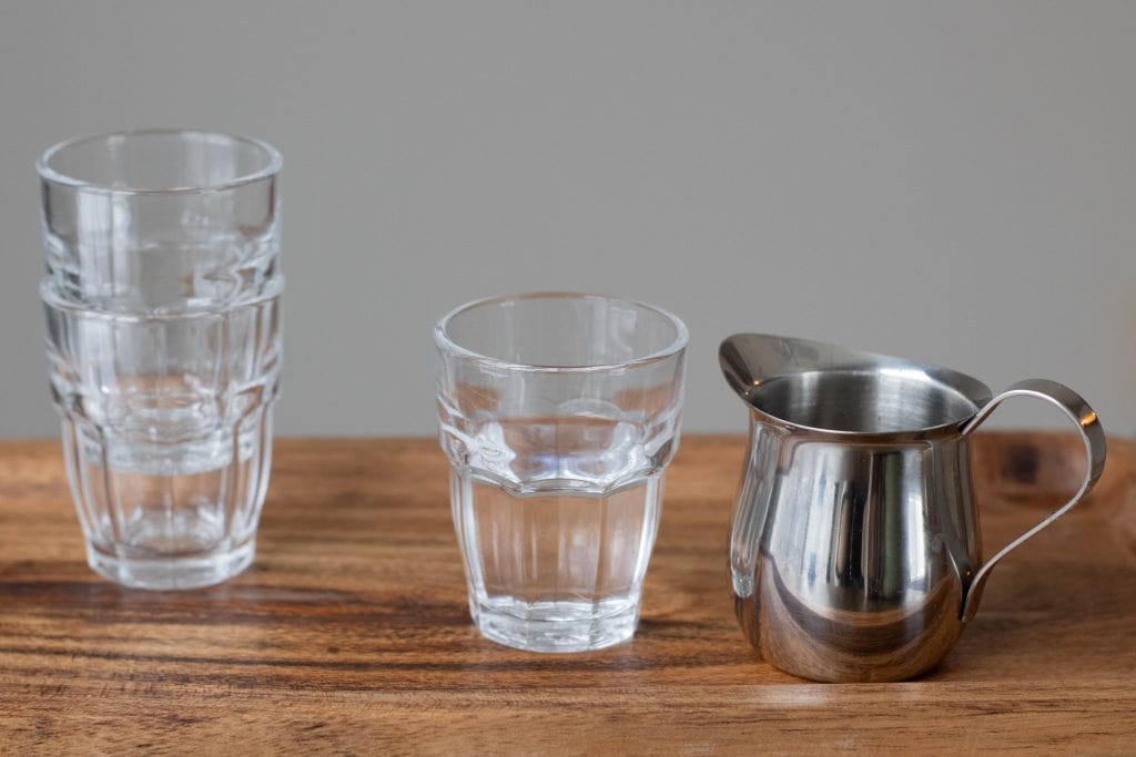 Two stacked glass cups one class cup half full with water and a small stainless steel pitcher. 