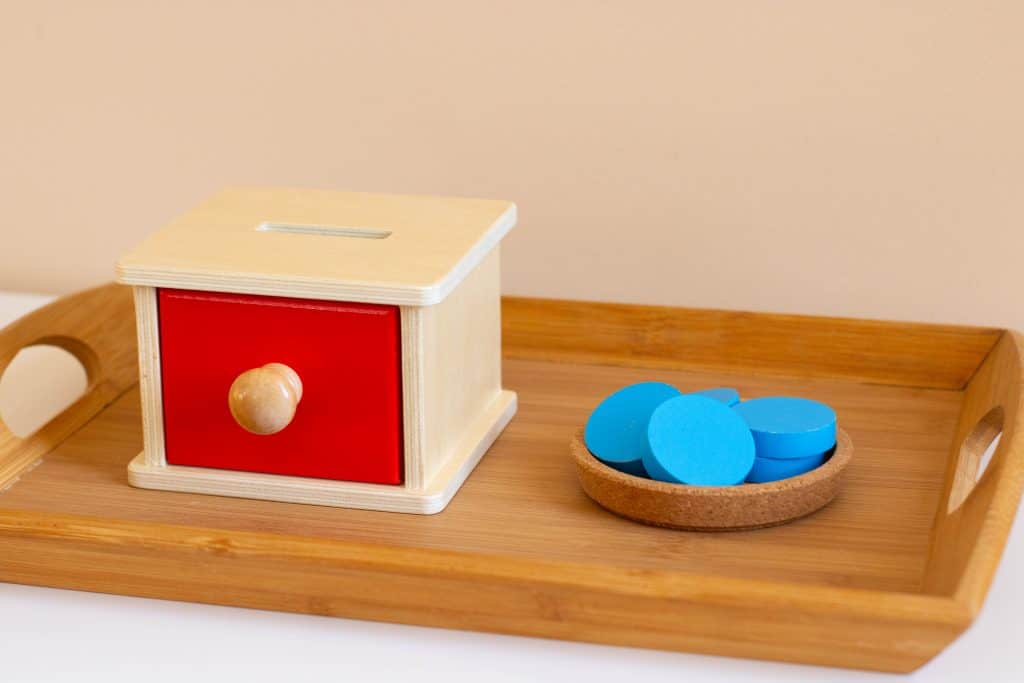 A tray with a wooden box that has a drawer with a wooden knob. A slit in the top of the wooden box and a dish of wooden coins.