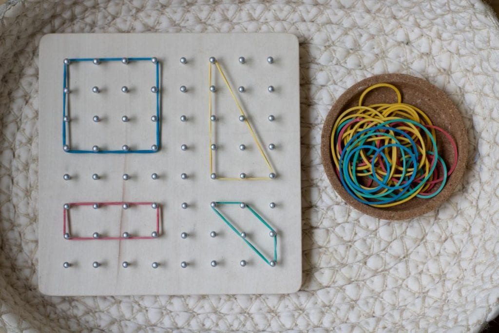 A wooden board of pegs with rubber bands stretched into shapes on the board.