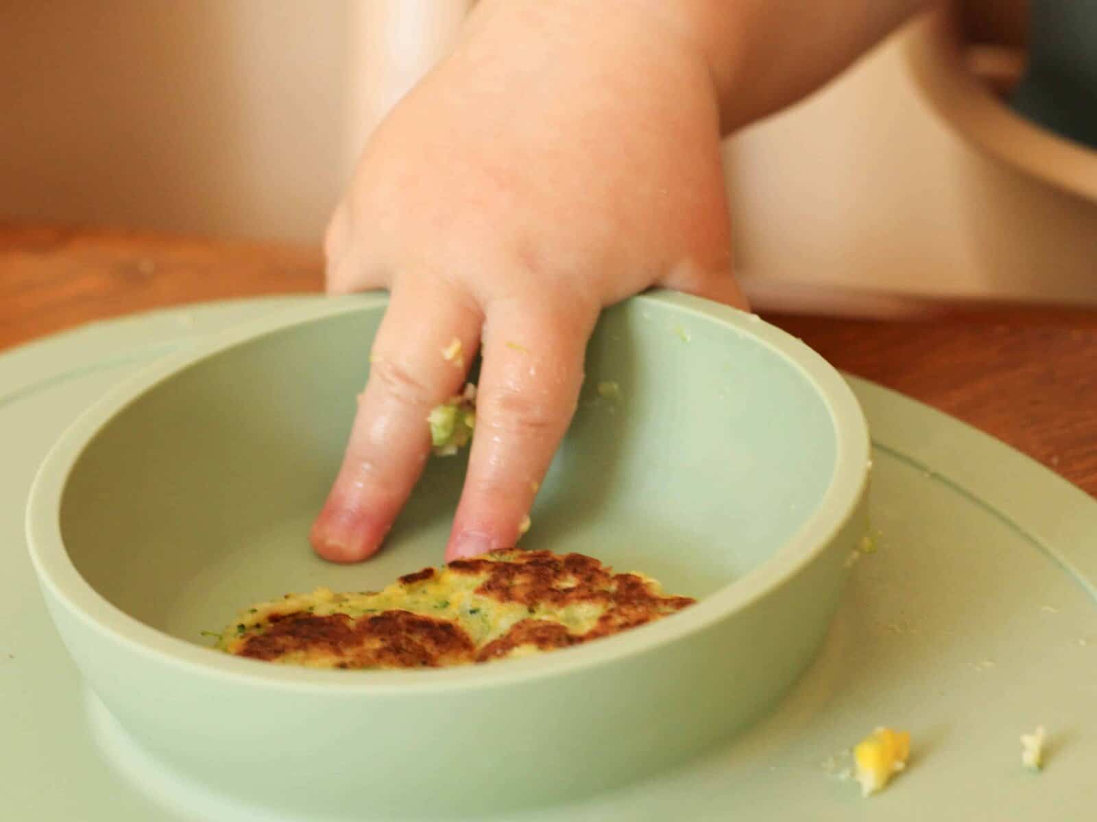 Close-up of a baby's hand reaching into a bowl to grab a sweetcorn fritter.