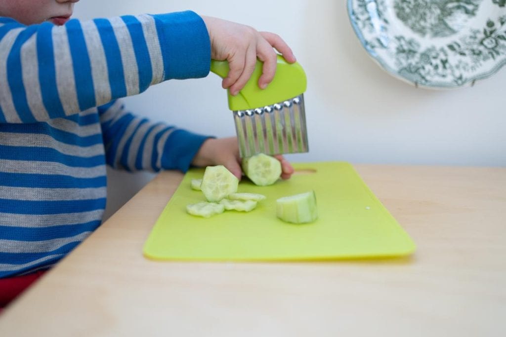 Child chopping a cucumber with a crinkle cutter.