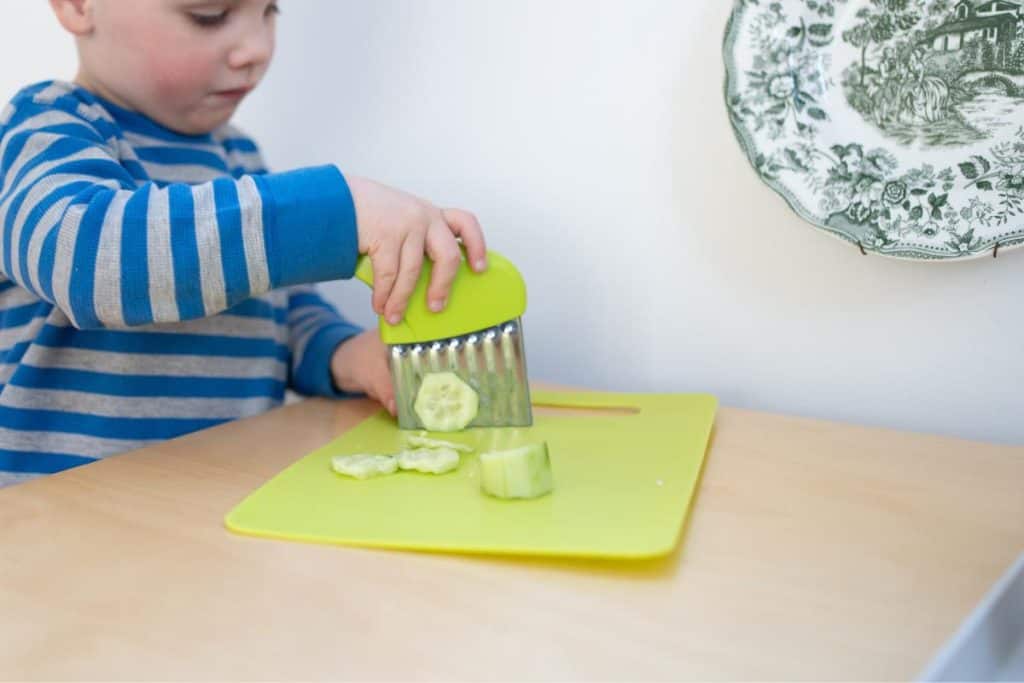 Child chopping a cucumber with a crinkle cutter.