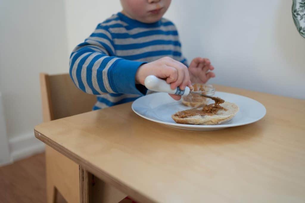 Child spreading peanut butter onto toast while sitting at a table.